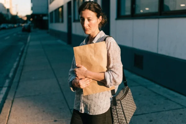 Mulher segurando um envelope pardo na calcada em frente a um predio comercial, no fim da tarde