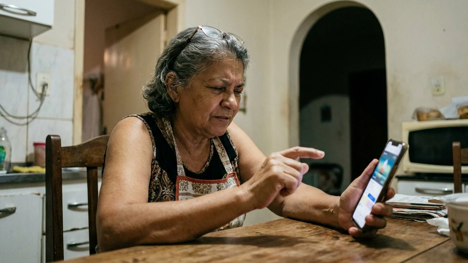 Mulher mais velha sentada à mesa da cozinha segurando o celular e hesitando antes de tocar na tela